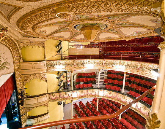 View of historic theatre auditorium from the upper circle, showing the ornate ceiling, top of the curtain above the stage and rows of seating.